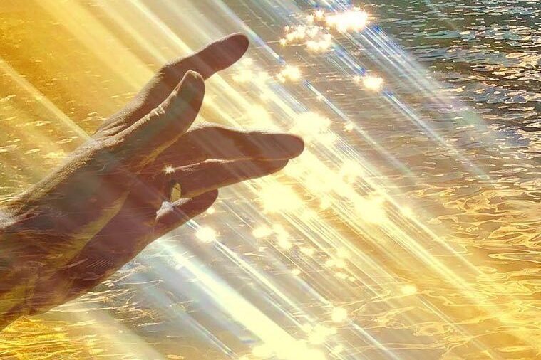 Image of a hand touching a beam of light with light sparkles falling into the ocean evoking the way Divine Spirit expresses Itself in the world as Light and Sound and the spiritual experiences we can have on our spiritual journey